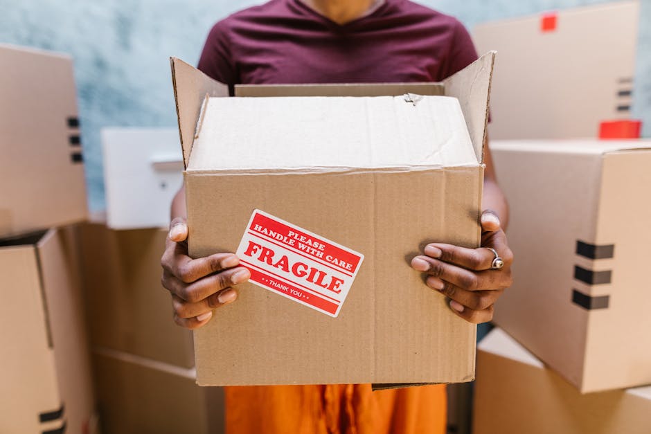 A scene of a home relocation process showing a room with cardboard moving boxes of various sizes stacked on the floor, some wrapped in plastic and others open with packing materials visible, positione